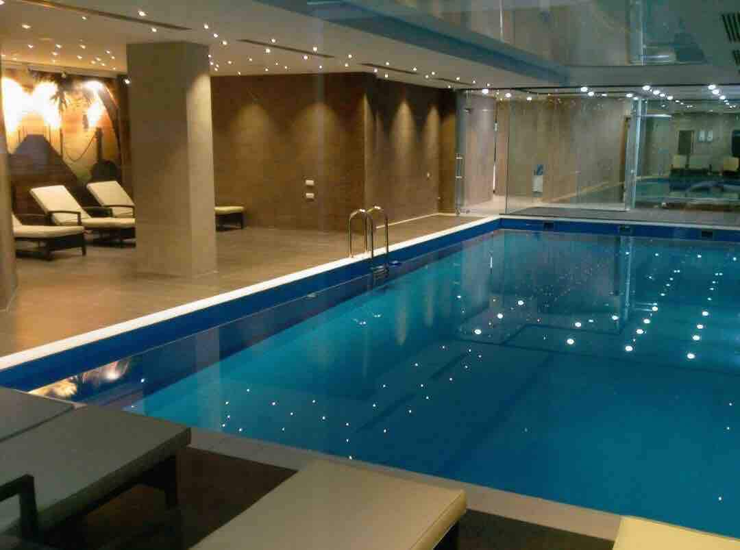 An indoor swimming pool is featured, surrounded by a clean, modern design. Lounge chairs are positioned along the edge, with soft lighting enhancing the tranquil ambiance. The water is a vibrant blue, reflecting lights from the ceiling above.