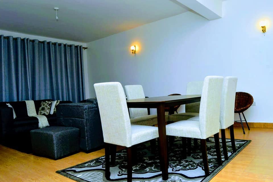 A modern dining area is highlighted by a dark wooden table surrounded by six light-colored chairs. A cozy seating arrangement is present nearby, complemented by a decorative rug and soft lighting. Floor-to-ceiling curtains add an element of warmth to the space.