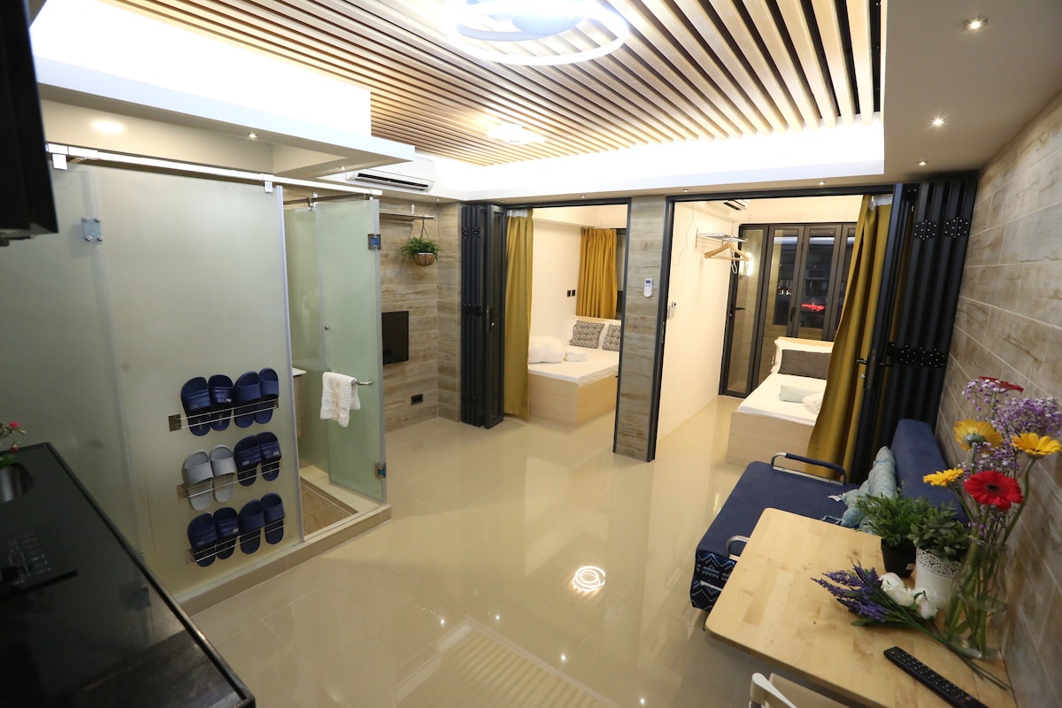 An open living space showcases a modern design, featuring a kitchenette with sleek cabinetry. A comfortable sofa faces a small dining area with a table. Two beds are visible in separate sections, and large glass panels provide natural light, complemented by warm wood accents and stylish decor.