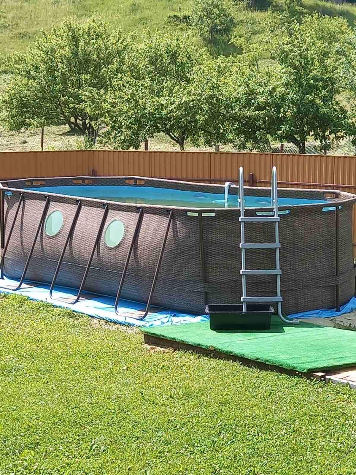 An above-ground pool is surrounded by lush green grass and a wooden fence. A metal ladder leads into the pool, which features circular windows along its sides. The pool area includes a safety mat on one side, creating an accessible entry point.