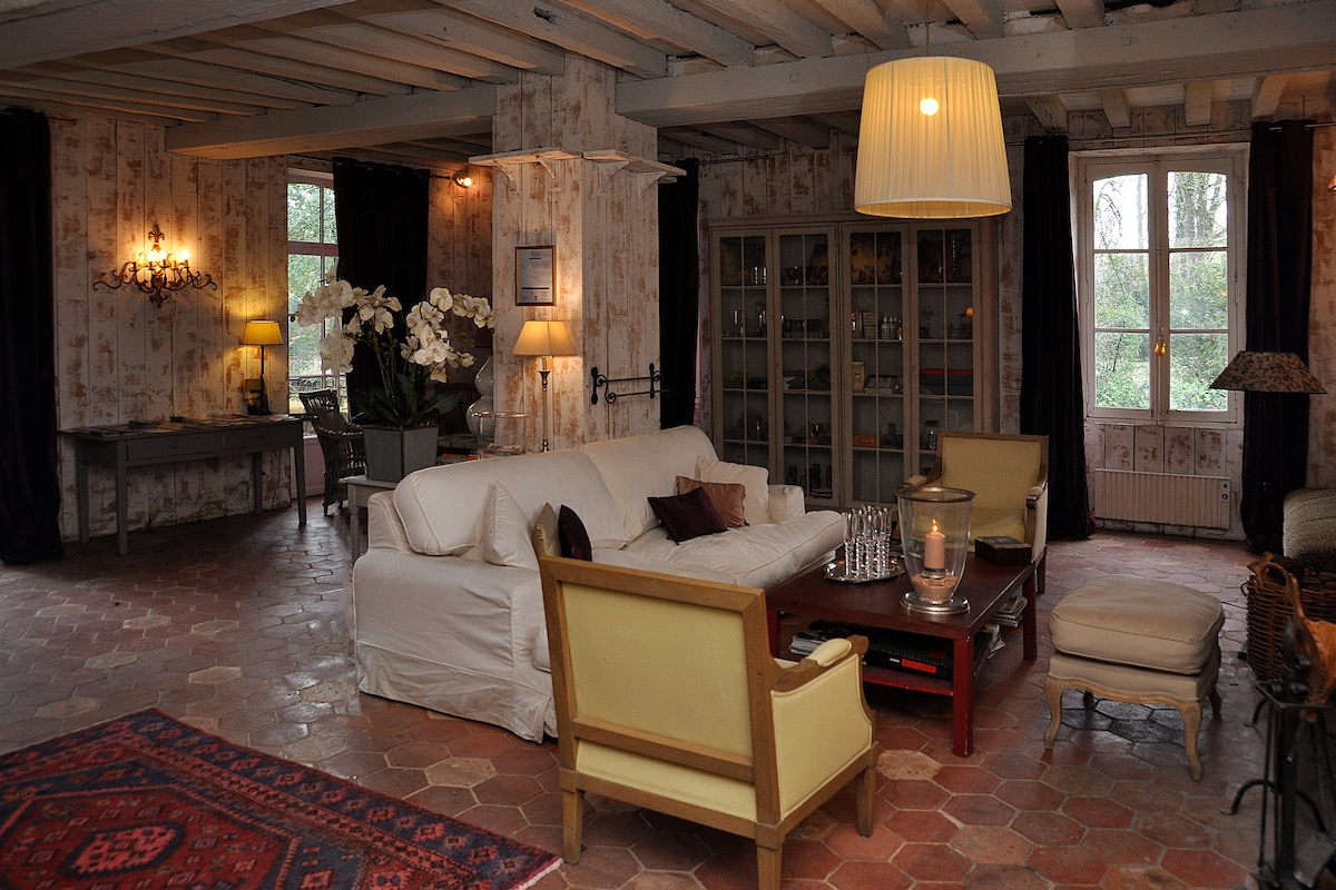 A spacious living area is highlighted by soft lighting and a warm color palette. A large white sofa and two yellow armchairs are arranged around a central coffee table. Decorative elements include a vintage rug and a display cabinet filled with glassware, complemented by large windows that allow natural light.