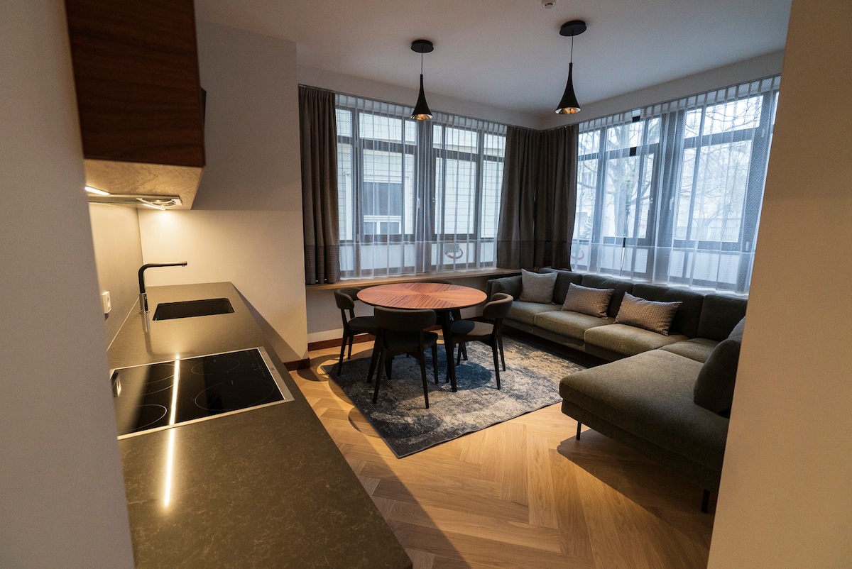 A modern living area is visible, featuring a round dining table surrounded by black chairs. A spacious sofa is positioned against the wall. Natural light filters through multiple large windows with sheer curtains, illuminating the stylish interior.
