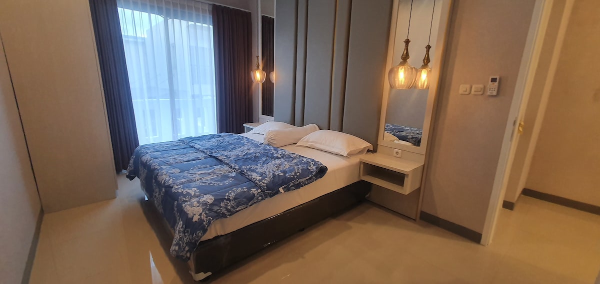A well-appointed bedroom features a spacious bed dressed in a blue floral comforter. Soft lighting from hanging pendant lamps creates a warm glow. Large windows are draped with sheer curtains, allowing natural light to filter into the space.