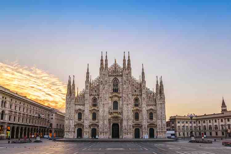 The stunning facade of a grand cathedral is showcased, featuring intricate spires and detailed stonework. The structure stands prominently in a wide square, surrounded by historic buildings under a vibrant sky during sunset.