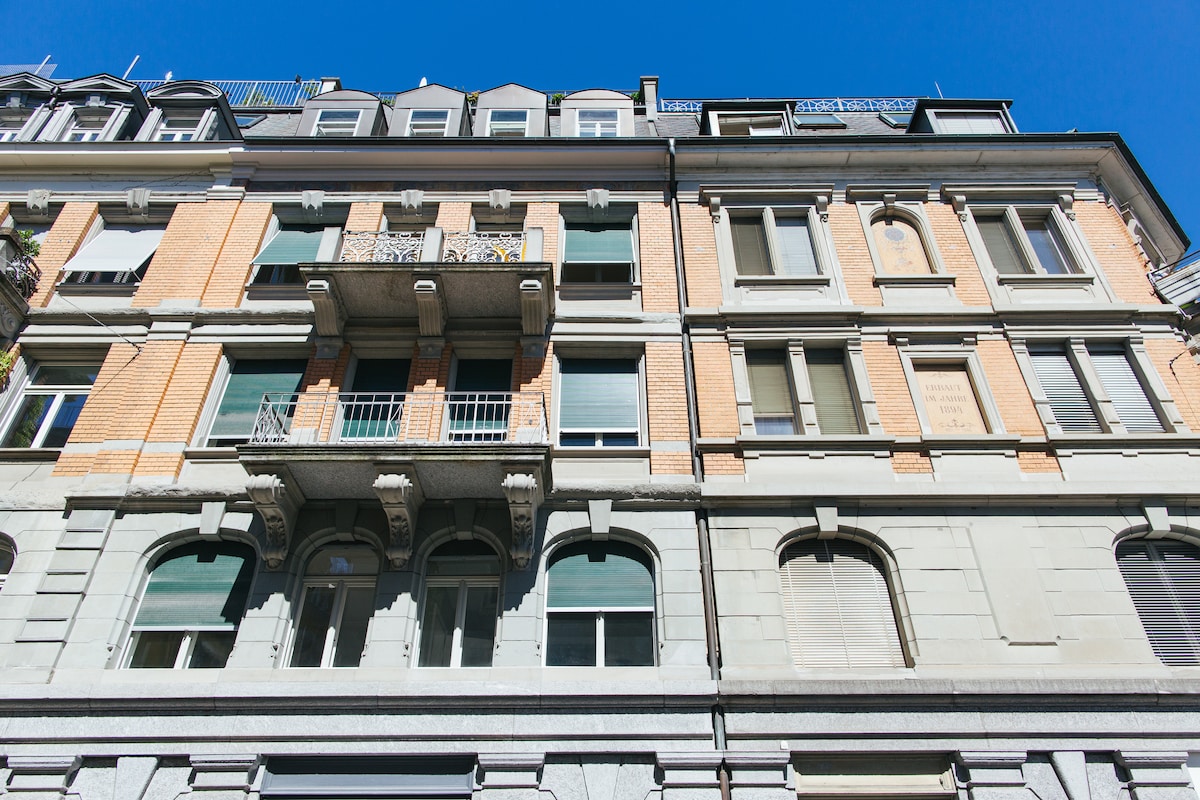 The historic facade of a renovated building is displayed, showcasing a blend of brick and stone elements. Decorative balconies can be seen on the upper floors, with large windows featuring shutters that allow natural light to filter in. The structure reflects architectural details typical of the late 19th century.