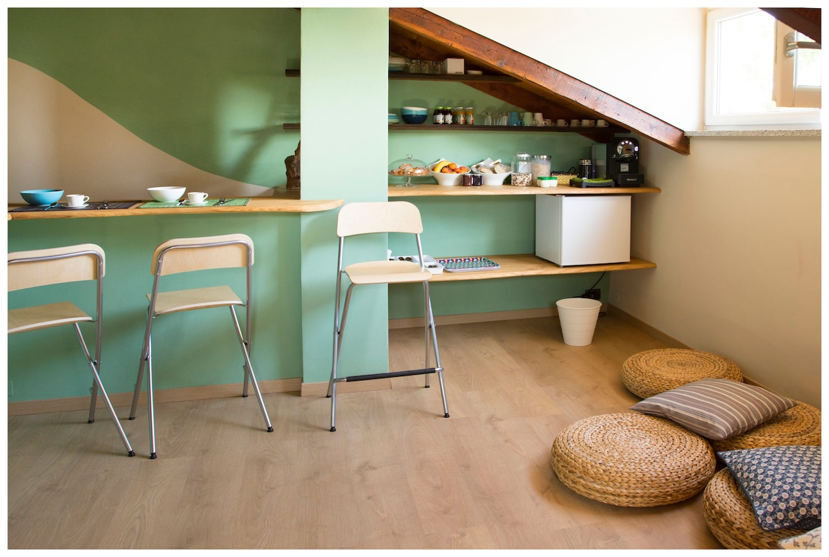 A spacious area features a green accent wall and wooden beams overhead. A bar counter with three chairs is present on one side, and a collection of breakfast items is arranged on shelves. Soft, round cushions are placed on the floor, inviting relaxation.