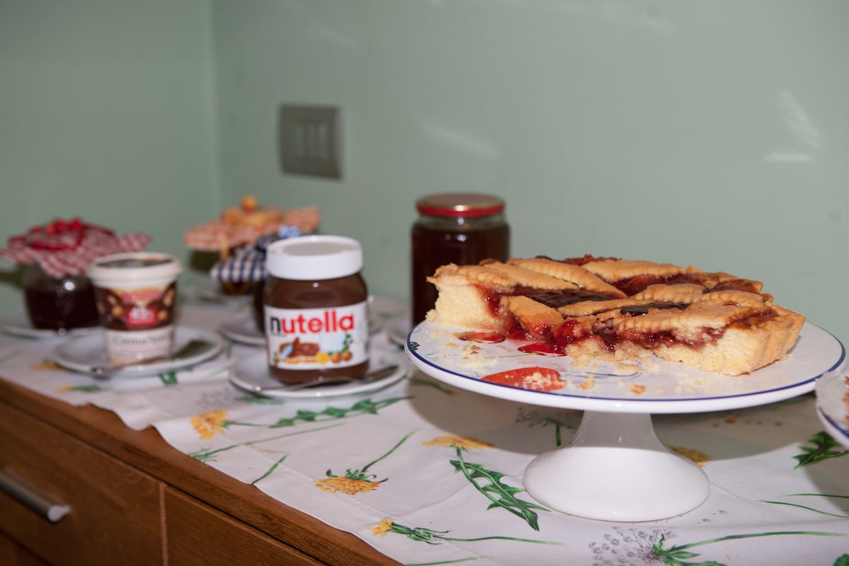A display of breakfast items is arranged on a table covered with a floral cloth. A slice of pie sits on a white stand, accompanied by jars of Nutella and assorted jams. Containers of yogurt are placed nearby, offering a variety of choices.