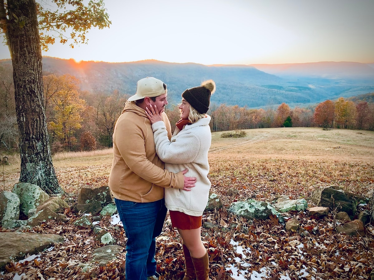 A couple stands close together on a grassy area surrounded by autumn foliage, with a sweeping view of rolling hills in the background. The sunset casts a warm glow across the landscape, enhancing the serene atmosphere.