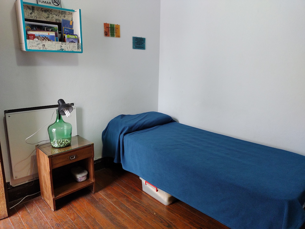 A cozy sleeping area features a single bed covered with a blue blanket. A bedside lamp is placed on a small wooden nightstand, while a wall-mounted shelf displays magazines and decorative items. Warm wood flooring adds to the inviting ambiance.