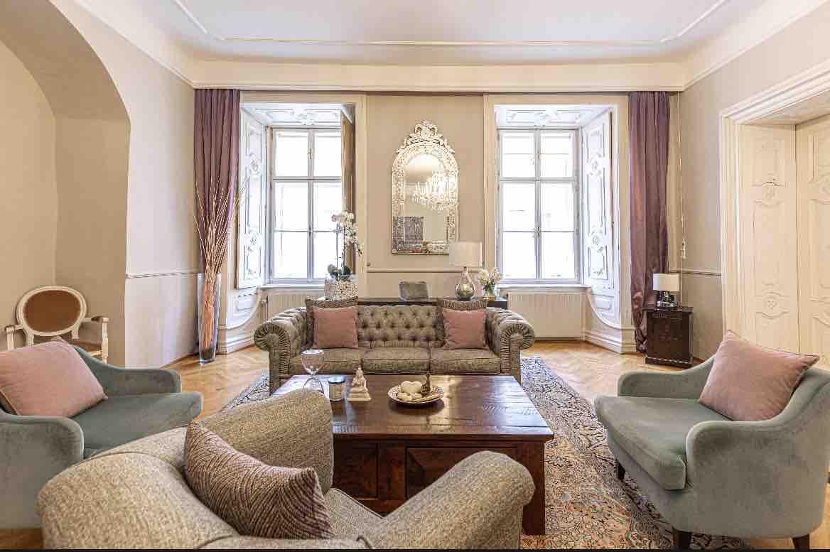 A spacious living room features a mix of upholstered seating with soft cushions and a wooden coffee table at its center. Two large windows allow natural light to illuminate the room, highlighting a decorative mirror and subtle wall decor. A warm, inviting atmosphere is present.