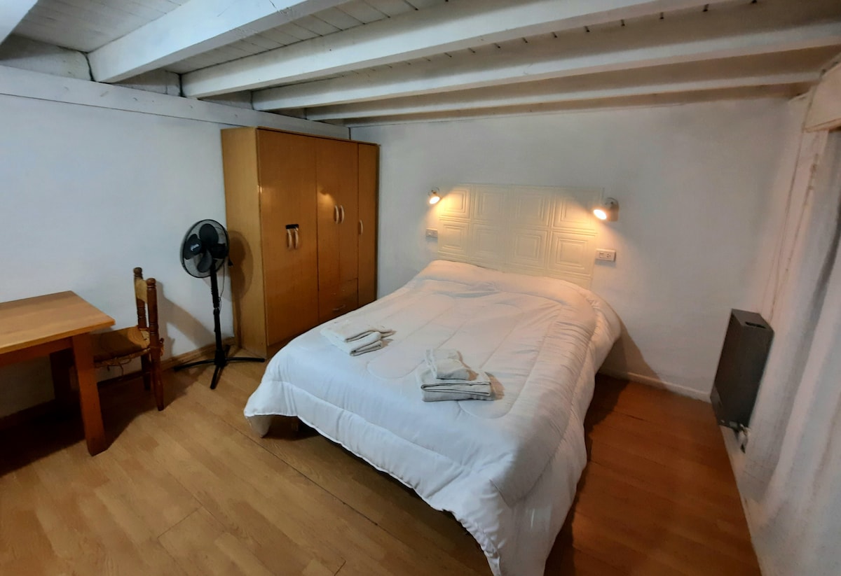 A simple bedroom features a spacious bed with a white comforter, complemented by two bedside lamps. A wooden table and chair sit near the bed, while a wardrobe stands against the wall. A fan and heater are also present, enhancing the functional layout.