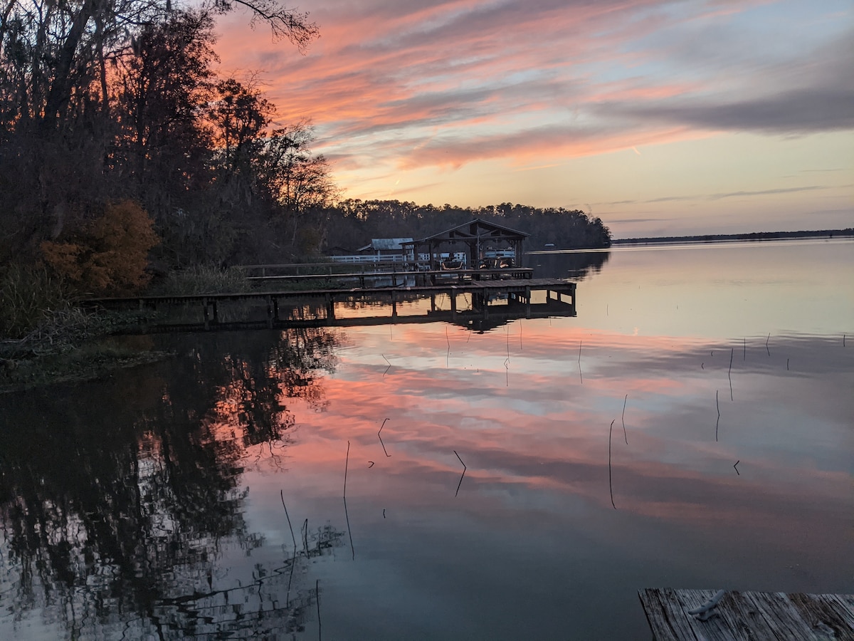 A serene view of Lake Seminole at sunset is captured, with reflections of pink and orange hues visible on the calm water. A wooden dock extends into the lake, framed by trees along the shoreline, creating a peaceful and scenic atmosphere.
