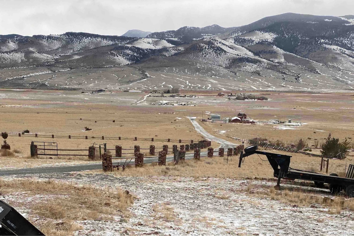 A wide view captures rolling hills and distant mountains dusted with snow. A gravel road meanders through an open landscape, flanked by barbed wire fencing. Scattered farm structures are visible in the valley, surrounded by natural scenery and gentle slopes.