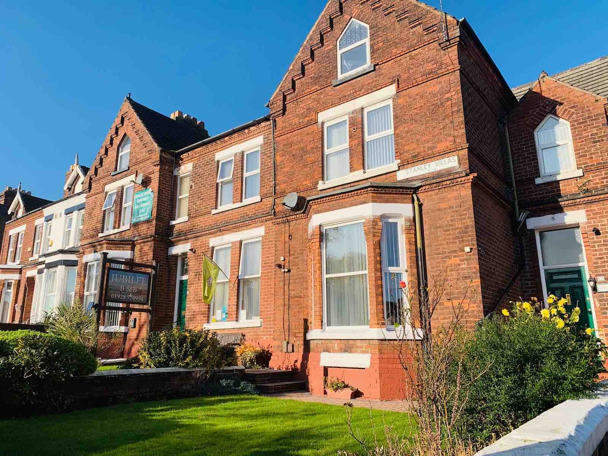 The exterior of a charming brick building is depicted, showcasing large windows and a well-maintained front lawn. A welcoming entrance is highlighted by the green canopy, while morning sunlight casts a warm glow on the facade. Neighboring structures add to the neighborhood's character.
