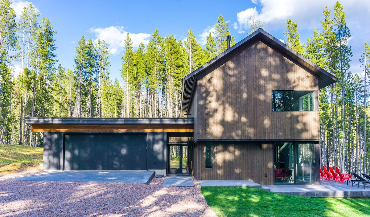 The exterior of the cabin showcases a modern design with a combination of dark wood and large glass windows. A spacious driveway leads to a two-car garage, while a natural landscape of tall trees surrounds the property, creating a tranquil setting.