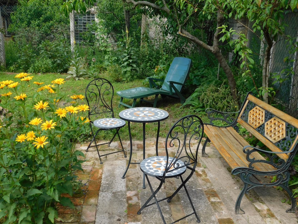 A peaceful garden area features a wooden bench and two small round tables with decorative tiles, surrounded by vibrant yellow flowers. A lounge chair is positioned nearby, with lush greenery providing a natural backdrop.