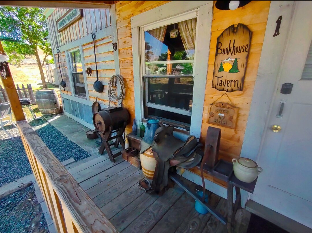 A rustic porch is visible, featuring a wooden sign that reads 'Bunkhouse Tavern.' Various vintage items, including a metal barrel, tools, and decorative containers, are arranged along the wooden decking, which is bordered by a pebbled pathway and greenery in the background.