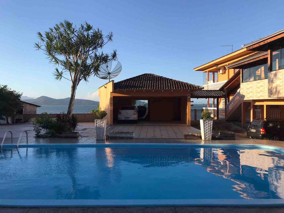 A large outdoor pool is framed by a tiled patio and potted plants, offering views of the surrounding landscape. A covered parking area is situated nearby, and a two-story building with a mix of wood and stone features stands in the background.