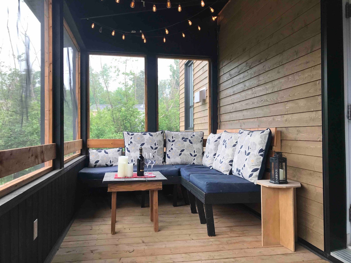 A screened-in porch is featured with a comfortable seating area, consisting of plush cushions and dark wood accents. A small wooden table rests at the center, accompanied by candles and a decorative lantern. Above, string lights create a soft glow suitable for relaxation.