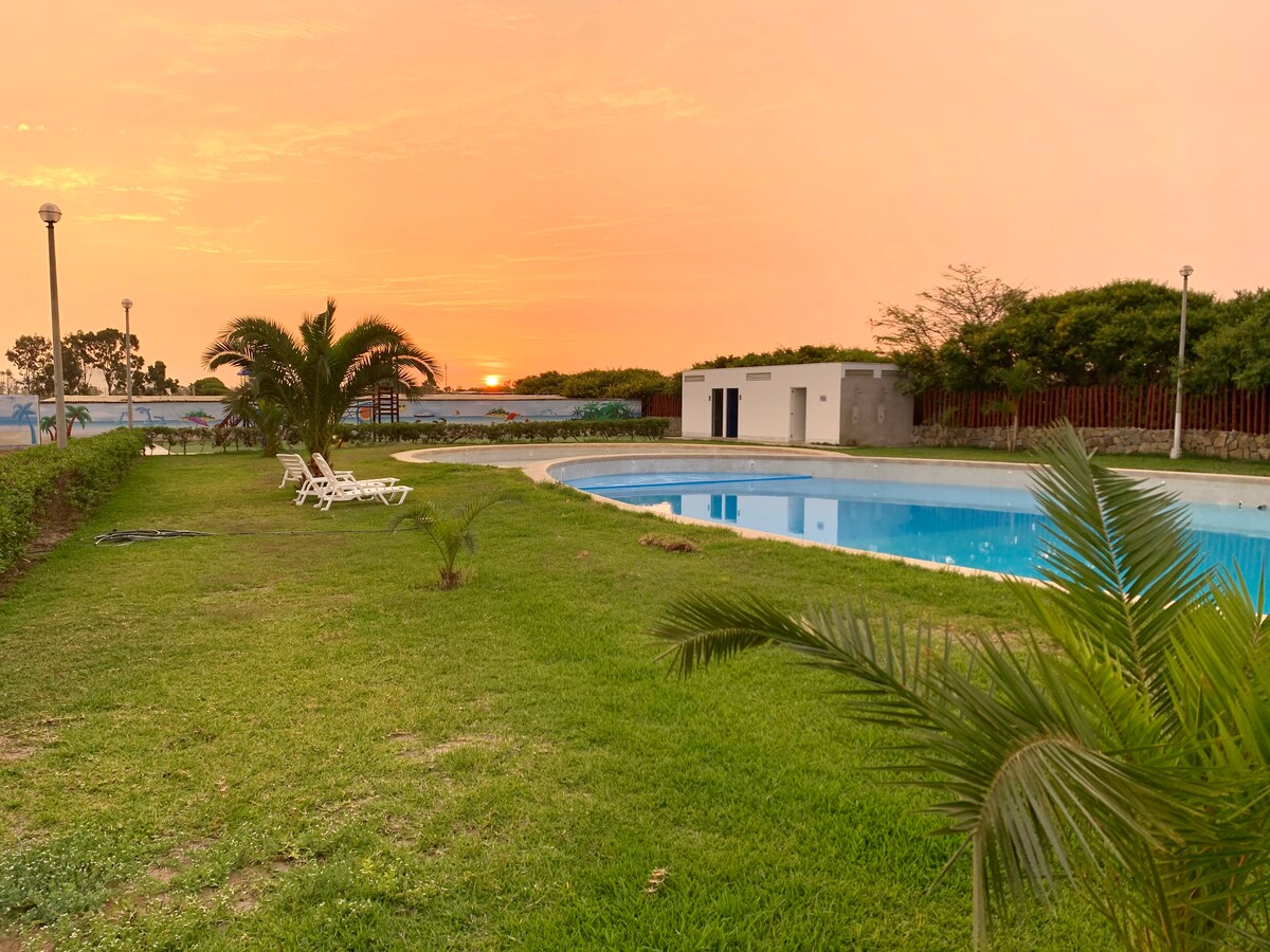 A serene pool area is surrounded by lush green grass and palm trees, with lounge chairs positioned for relaxation. In the background, a soft sunset casts a warm glow over the water, reflecting the evening sky.