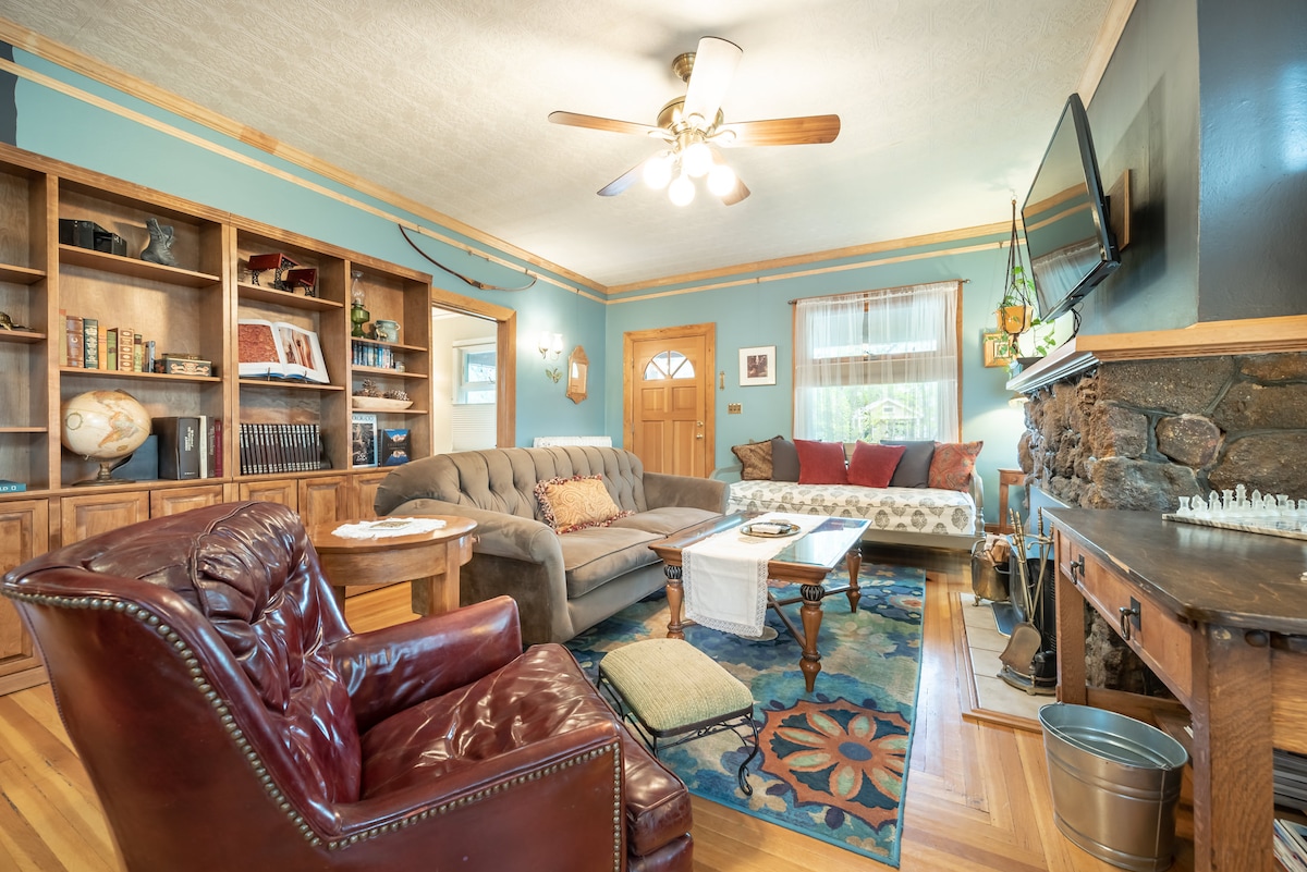 The living area features a soft sectional sofa complemented by a classic armchair. A bookshelf lines the wall, showcasing various decor. Central lighting is provided by a ceiling fan, and a decorative area rug adds color to the hardwood floor. A mounted TV is present above a traditional fireplace.