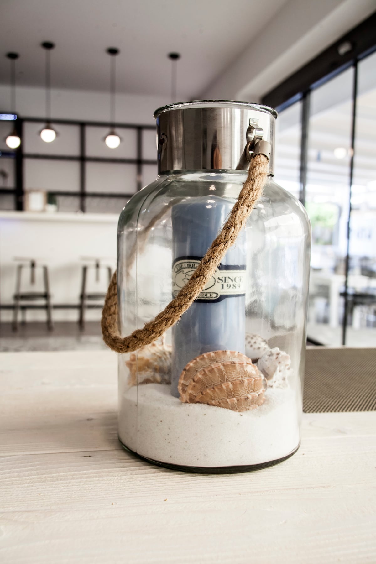 A glass jar with a metal lid and a jute handle contains white sand and various seashells. A blue candle stands in the center, providing a coastal decorative touch. The background features a modern bar area with stools and bright lighting.