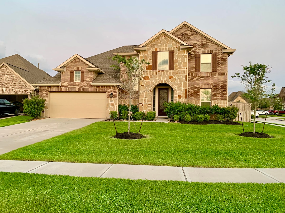 A spacious home is showcased with a mix of brick and stone exterior. A neatly manicured lawn surrounds the entry, enhanced by young shrubs and small trees. The driveway provides ample parking space, leading to an inviting front entrance.