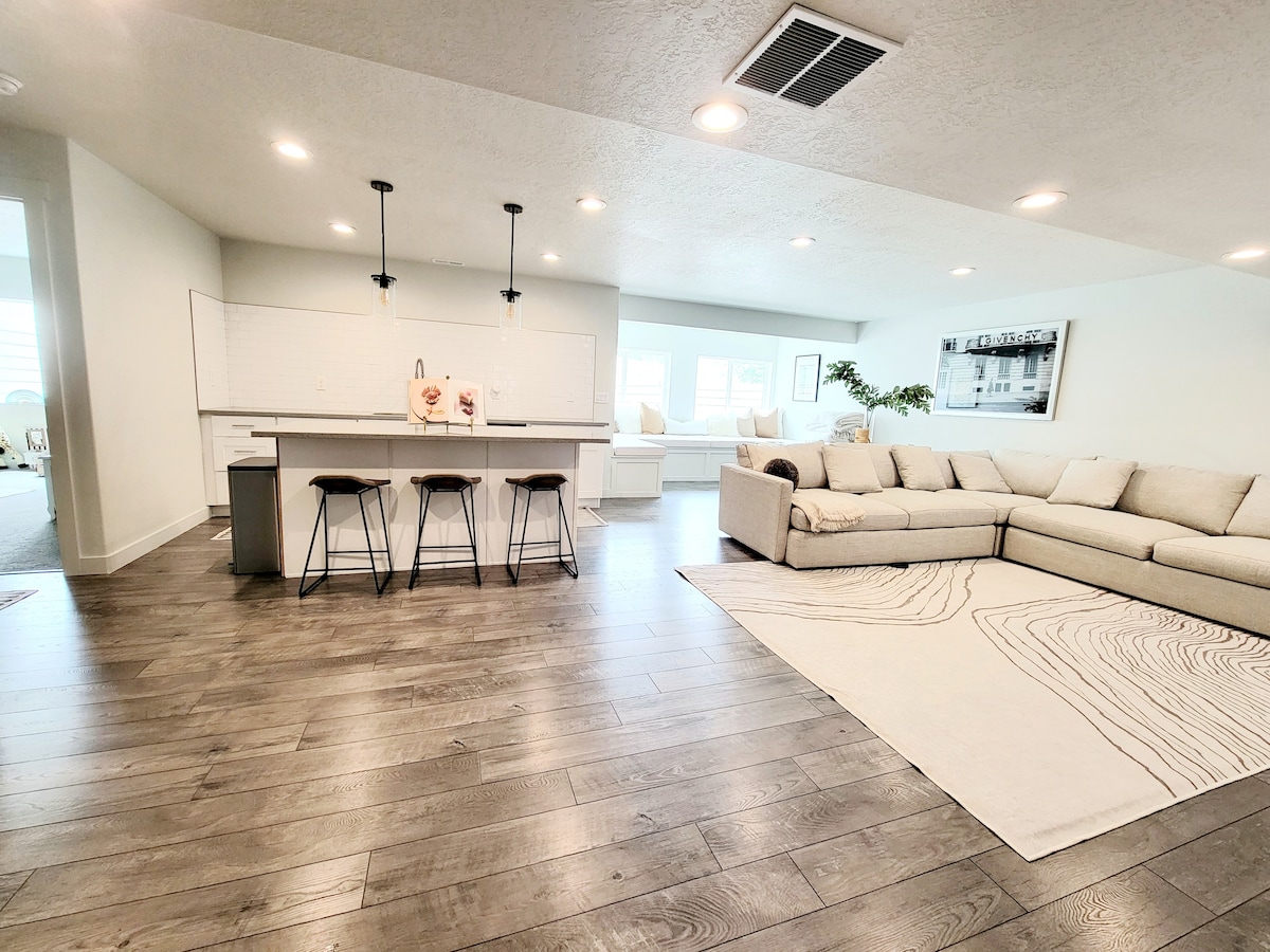 An open-concept living area features a modern dining space with three barstools positioned at a large kitchen island. A spacious sectional sofa sits on light wood flooring in the living room, which is filled with natural light from large windows.