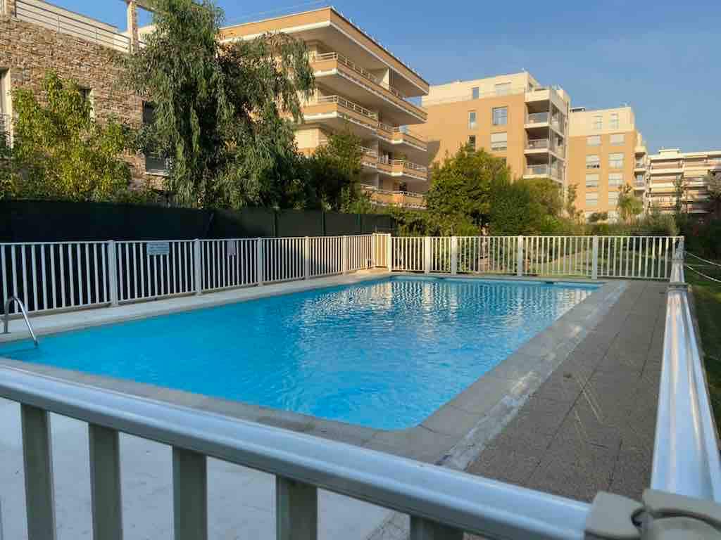 A securely fenced swimming pool is visible, featuring clear blue water under a bright sky. Surrounding greenery includes trees and shrubs, with apartment buildings visible in the background. The pool area is designed for relaxation and leisure.