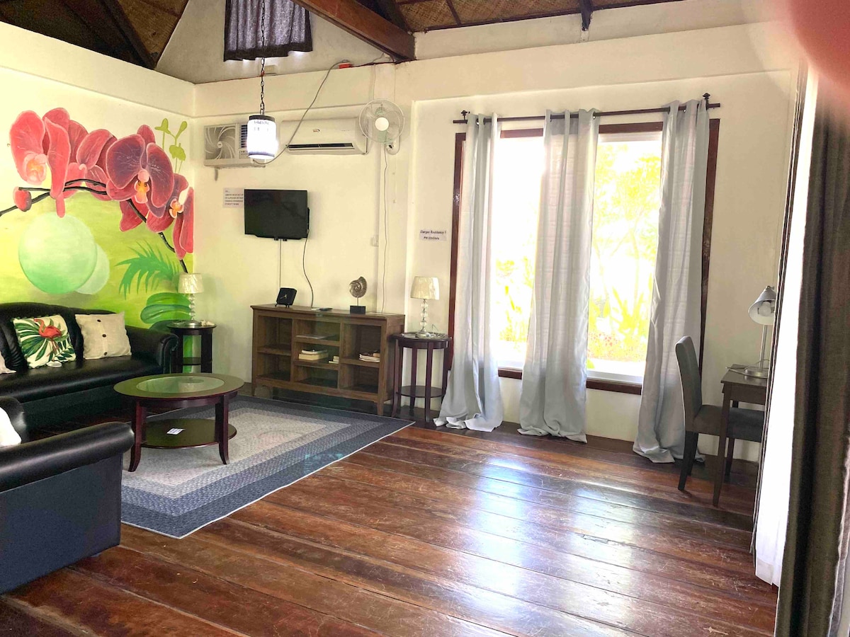 A living area is showcased with dark wood flooring and white walls. A vibrant mural of orchids adds color to the space. A comfortable couch is positioned near a coffee table, while a small desk and lamp are visible. Light filters through the tall, sheer curtains at the window.