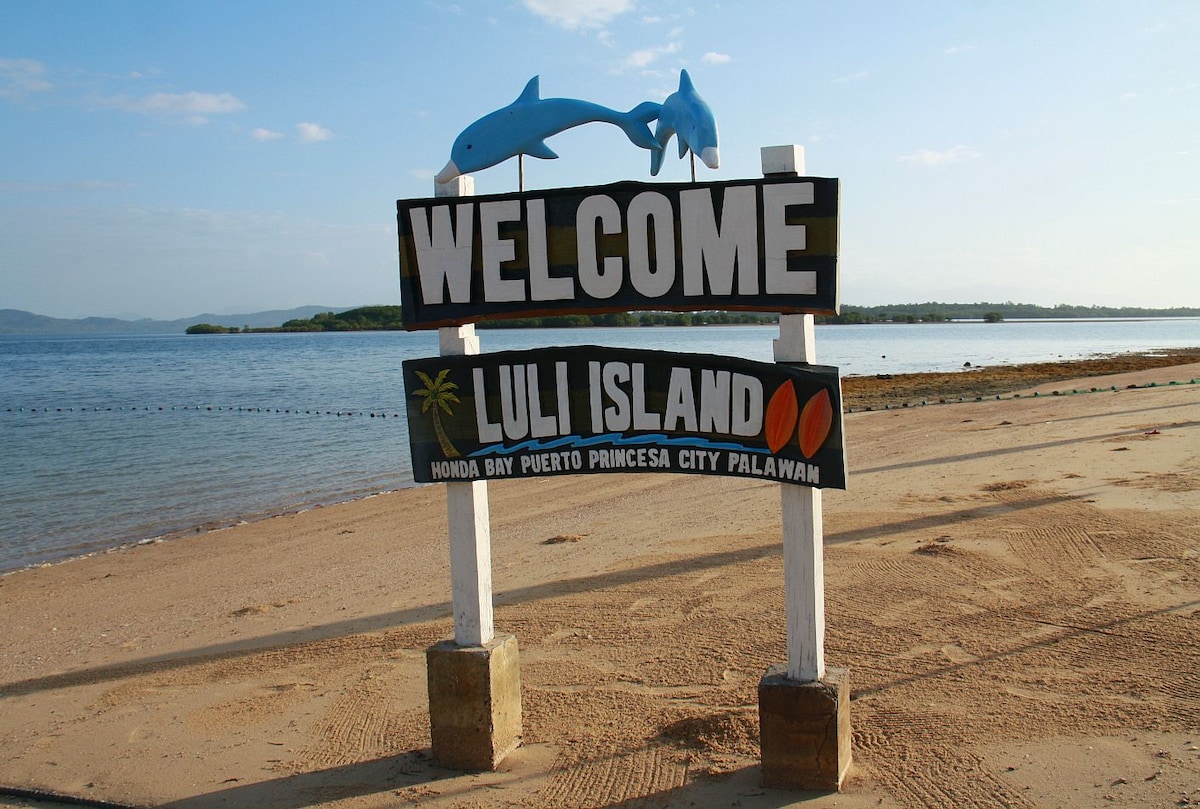 A welcoming sign for Luli Island is prominently displayed against a backdrop of the calm sea and a clear blue sky. The sign features vibrant colors, including images of dolphins and tropical elements, positioned on sandy terrain that leads to the water's edge.