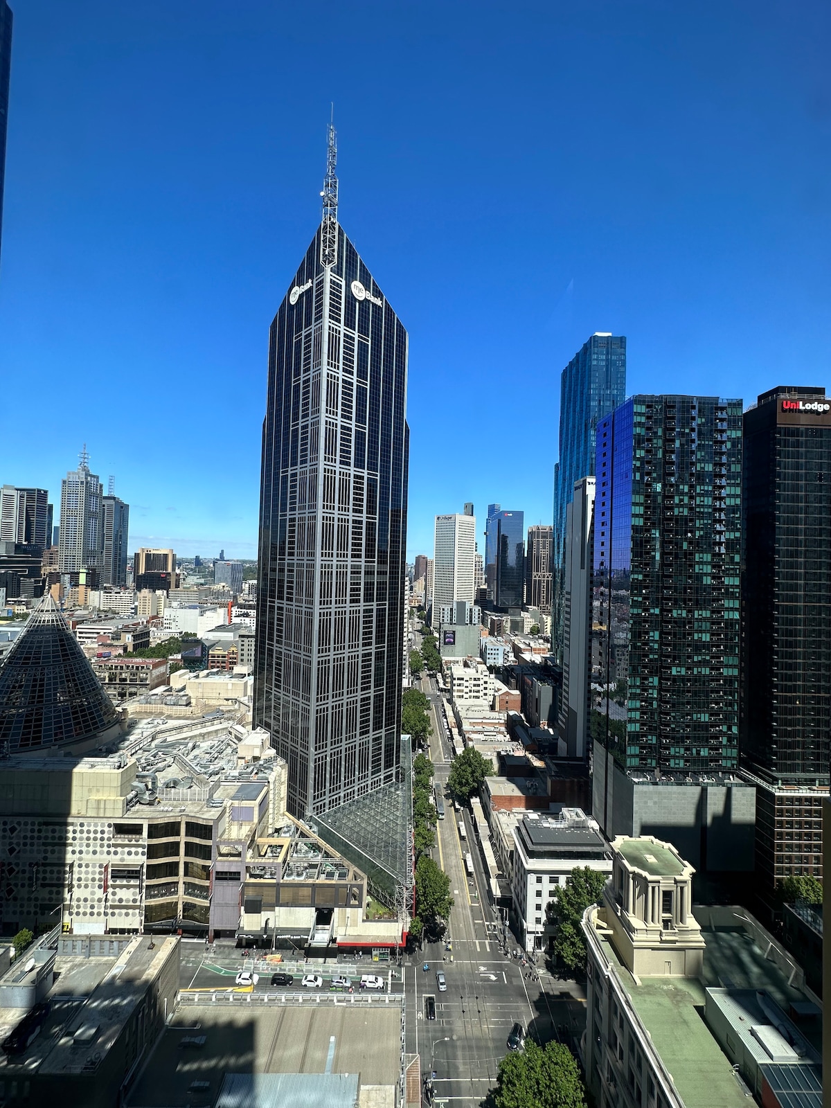 A view of the Melbourne skyline features a prominent tall building with a unique design, surrounded by a mix of modern skyscrapers and city structures. Clear blue skies overhead complement the urban landscape, showcasing a vibrant city atmosphere.