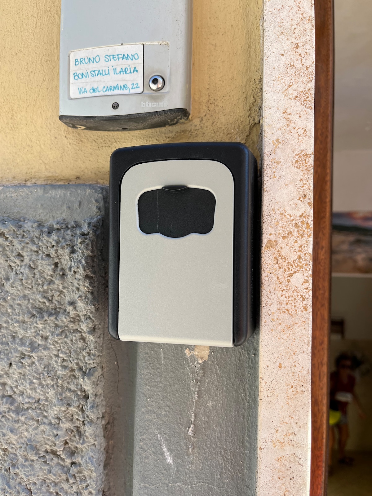 A mounted key box is positioned on a wall near a door, featuring a black and gray exterior. The box's front displays a slot for depositing keys, with a label attached above it. Surrounding surfaces include textured stone and painted wall elements.