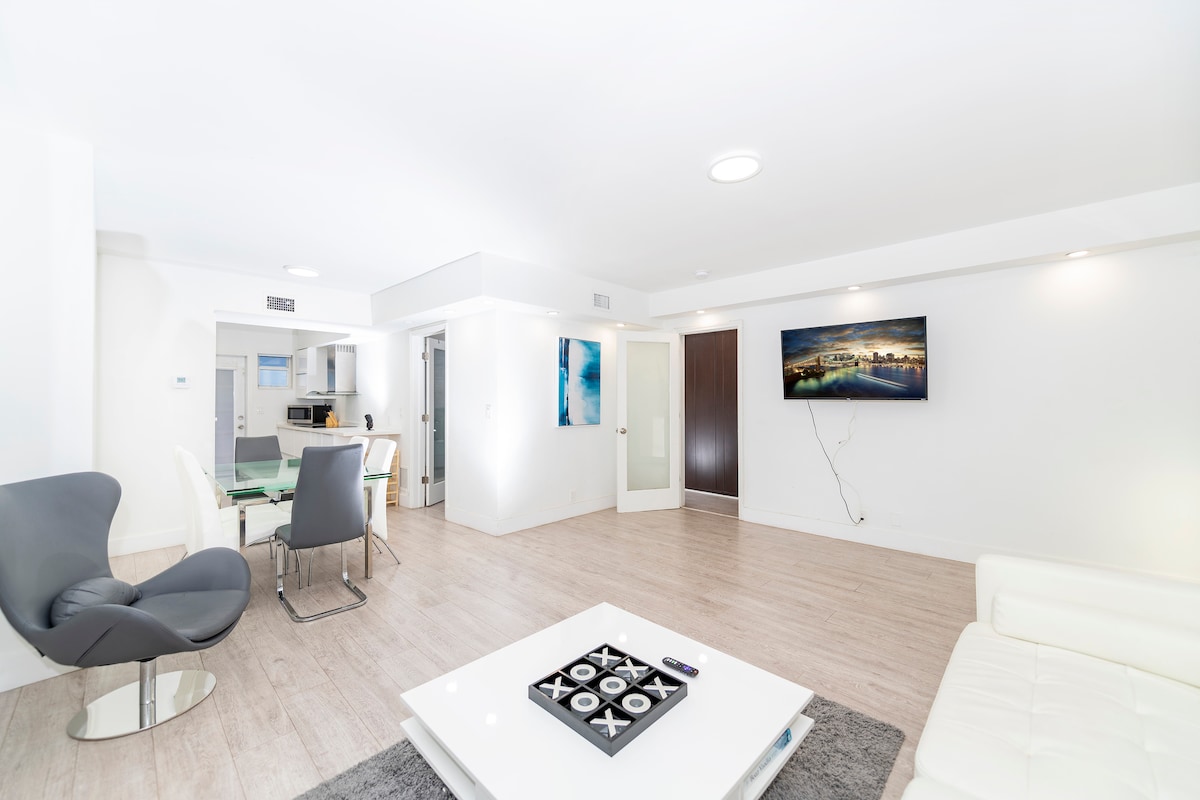 A modern living area features light-colored wood flooring and a white leather sofa. A sleek coffee table sits in the center, while a flat-screen TV is mounted on the wall. A dining area with a glass table and chairs is visible in the background.