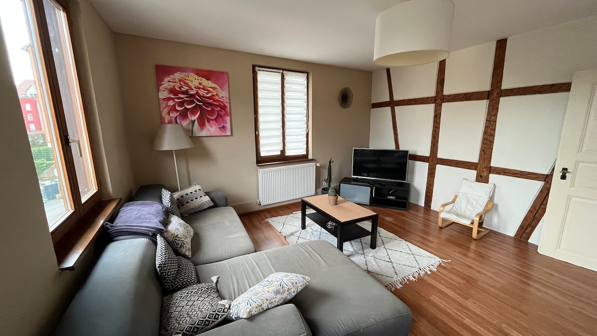 A well-furnished living room features a gray sectional sofa, a wooden coffee table, and a small armchair. Natural light fills the space through two windows with wooden shutters. A wall with exposed beams adds character, while a vibrant floral artwork decorates one wall.