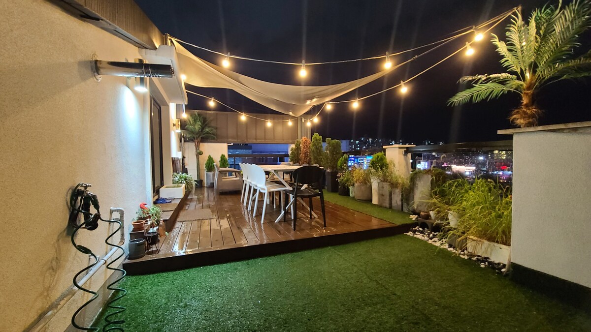 A rooftop terrace is illuminated by string lights, creating a cozy atmosphere. The wooden deck features white chairs and a table, surrounded by potted plants and ornamental grasses. A section of artificial grass adds green to the space, with a view of the city skyline in the background.