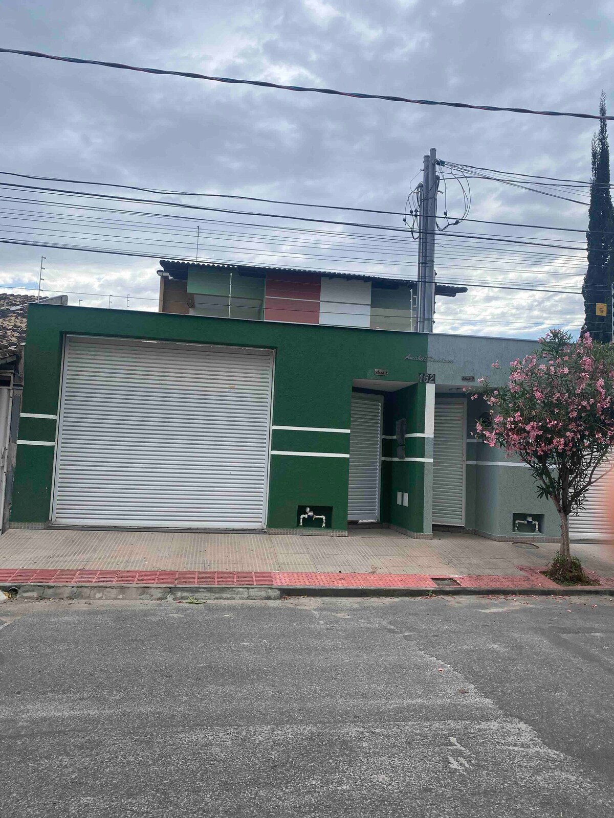 A modern two-story structure is portrayed, featuring a bold green exterior with white stripes. The entrance is accessible through a door beside a large garage door. Surrounding the building are minimal landscaping elements and a flowering tree, set against a cloudy sky.