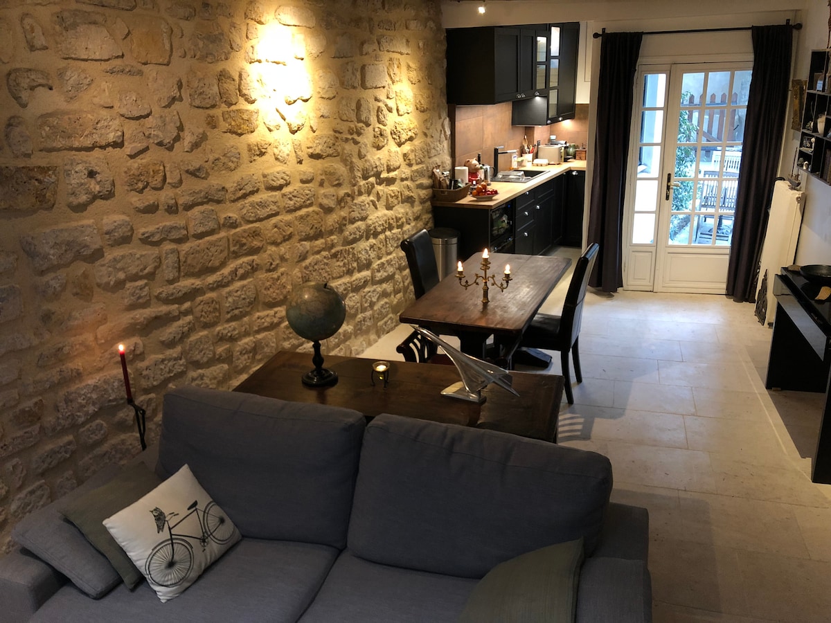 The image showcases a combined living and dining area with a stone wall feature. A comfortable sofa is positioned toward a dining table set for four. The kitchen is visible in the background, featuring dark cabinetry. Light enters through French doors leading to an outdoor space.