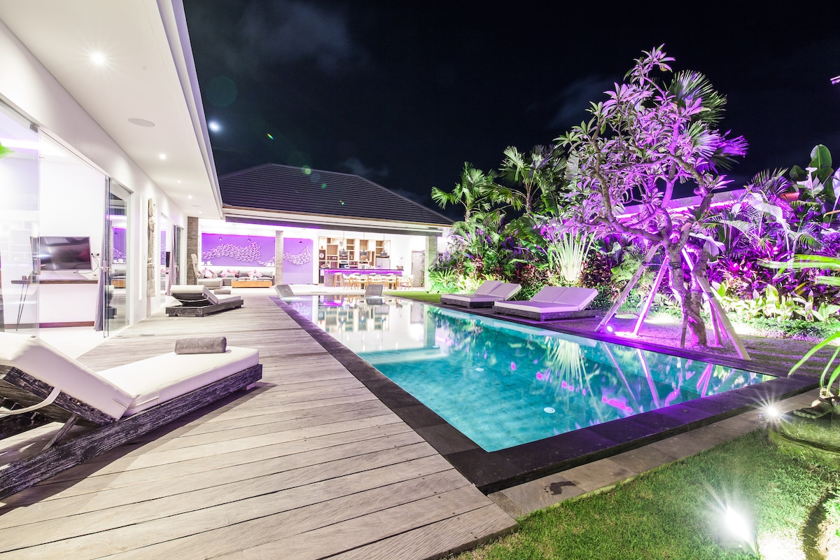 The tranquil pool area is illuminated by soft purple lighting at night, surrounded by lush greenery. Comfortable lounge chairs are positioned on a wooden deck, inviting relaxation. The villa’s interior is partially visible, highlighting an open living space that connects with the outdoor ambiance.