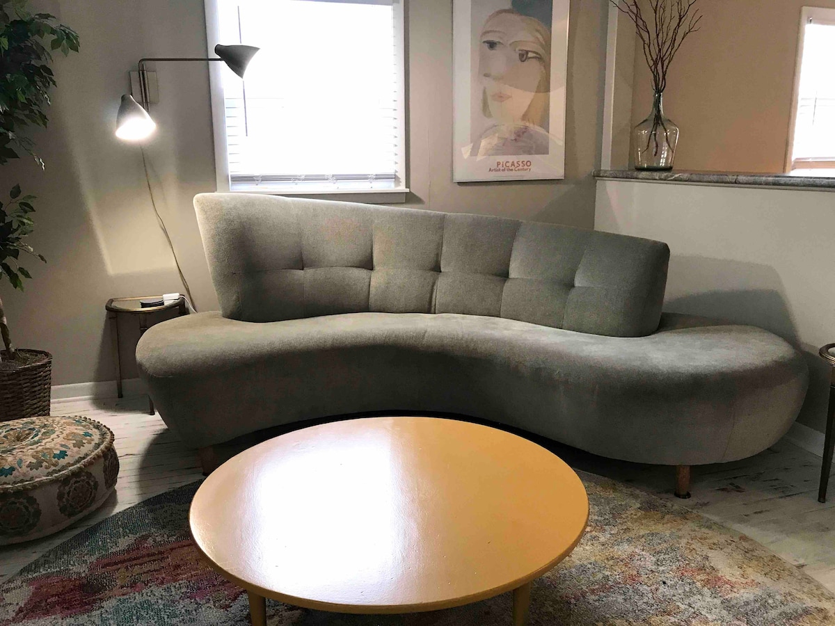A curved, soft sofa in a muted tone is positioned near a large window, providing ample natural light. A round yellow coffee table sits in front, and a patterned area rug adds warmth to the space. A sleek floor lamp stands beside the sofa.