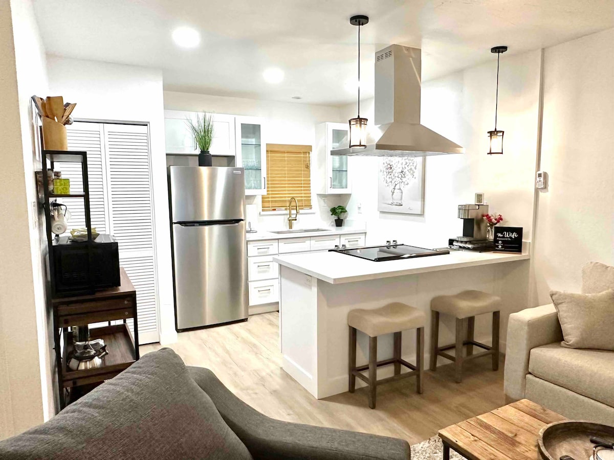 A modern kitchen area is featured, equipped with stainless steel appliances, including a refrigerator and a range hood. A spacious island with seating for two is present, alongside wooden cabinetry. The area is illuminated with pendant lights, creating a well-lit atmosphere.