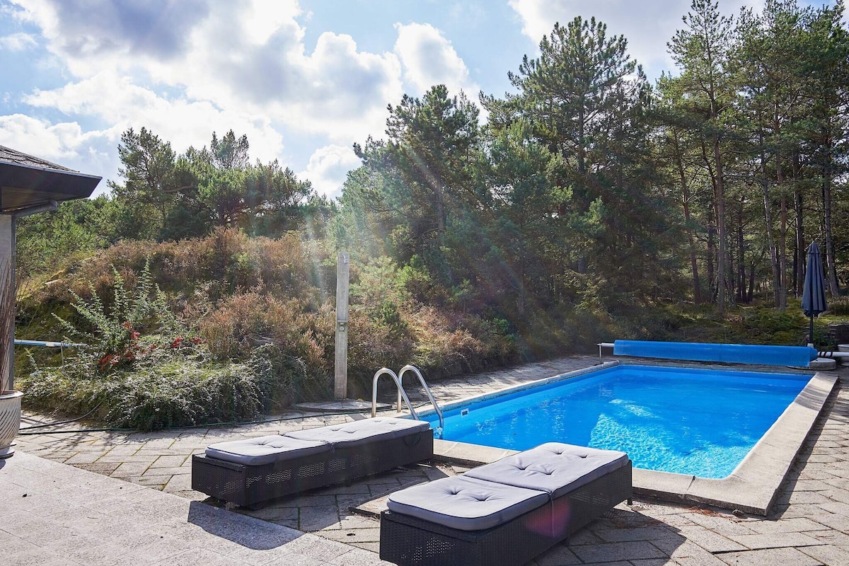 A heated outdoor pool is set against a backdrop of lush greenery and tall trees. Two lounge chairs are positioned beside the pool on a stone patio, providing a relaxing area for sunbathing and enjoying the natural surroundings.