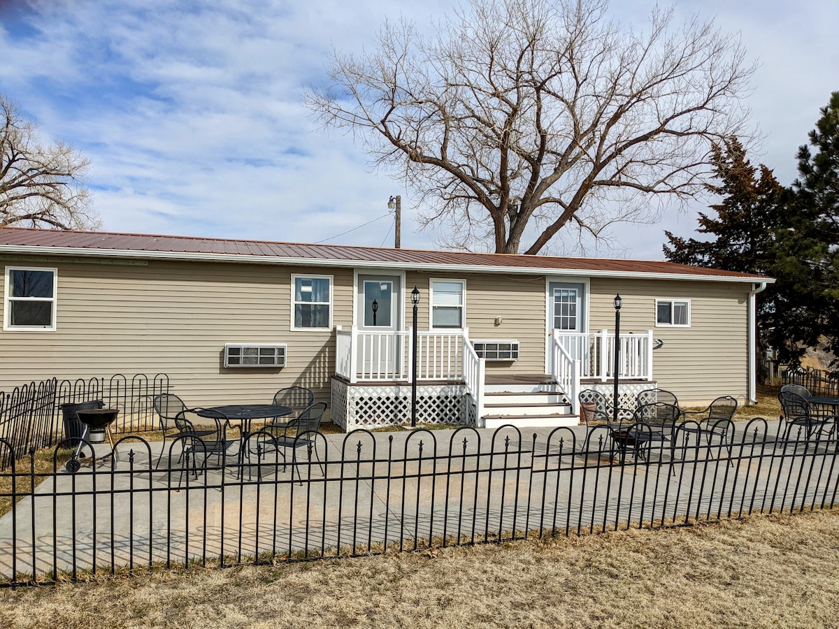 The exterior of a duplex modular home is shown, featuring a light-colored facade and a prominent entrance with steps. A fenced patio area hosts several black metal tables and chairs. Nearby, a tree provides shade, contributing to the calm surroundings.