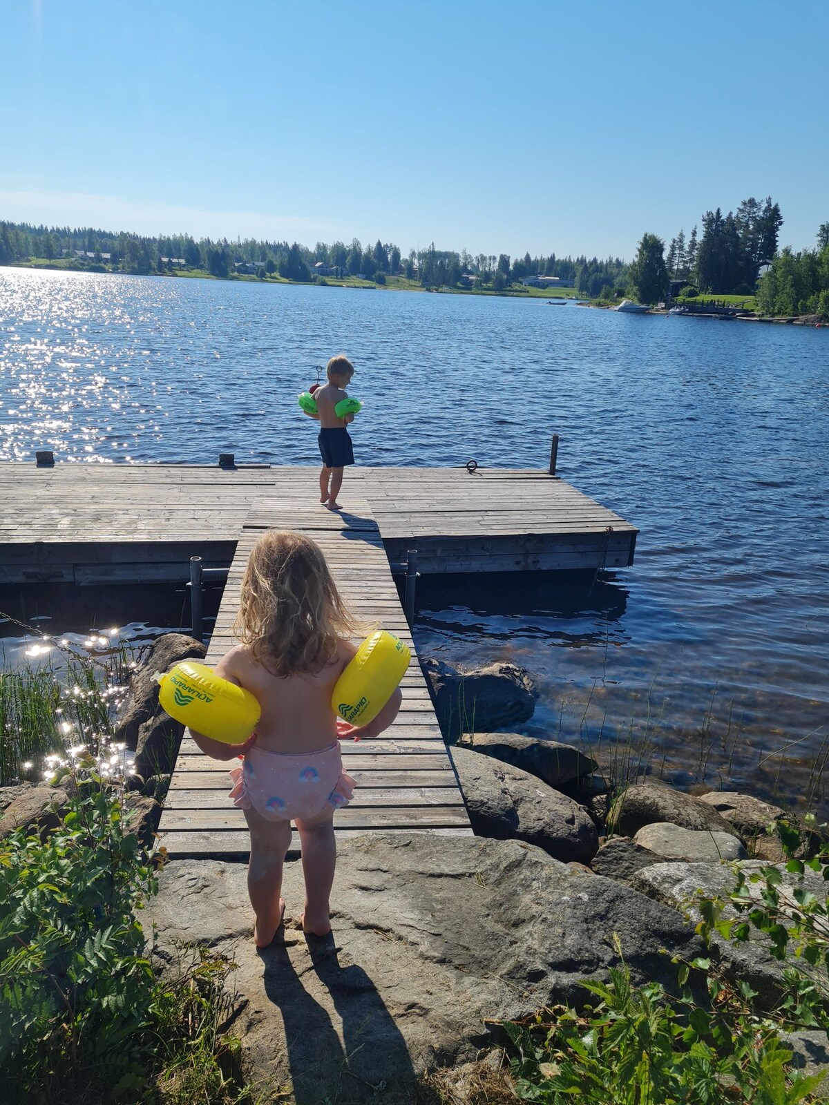 A wooden dock extends over calm water, surrounded by lush greenery. Two children are seen on the dock, one facing the water and the other turned slightly away. Sunlight glistens on the water's surface, creating a serene atmosphere.