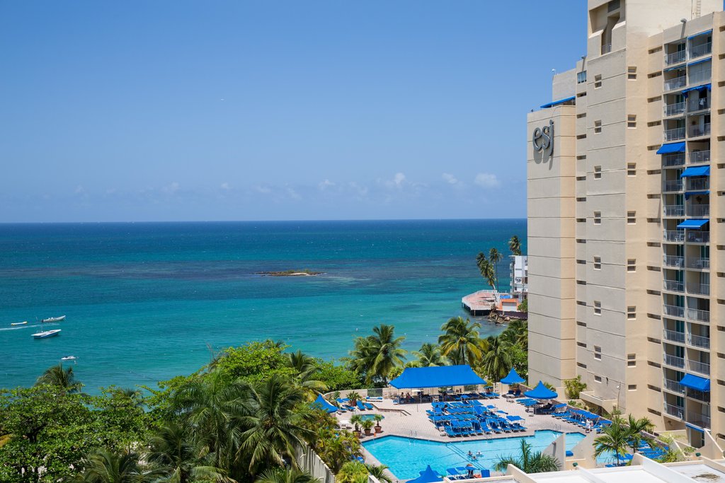 A view reveals the vast blue ocean extending toward the horizon, with small boats visible on the water. Tropical vegetation frames the scene, while a pool area, surrounded by sun loungers and umbrellas, is set below the hotel, enhancing the coastal landscape.
