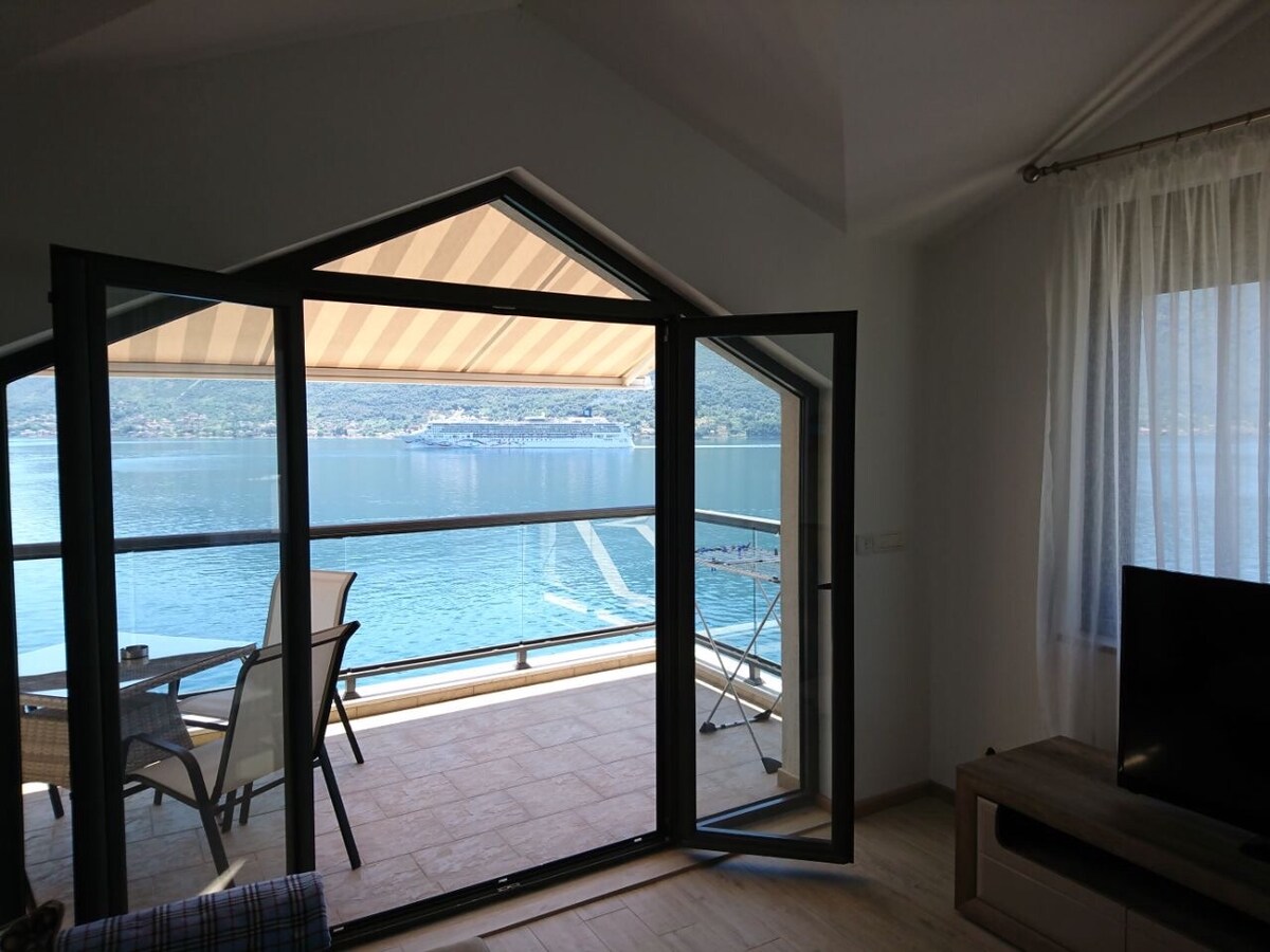 Large glass doors open to a balcony overlooking a serene body of water. A dining table with chairs is positioned on the balcony, while natural light fills the bright living space. The room features a TV and light-colored furnishings, enhancing the relaxing atmosphere.