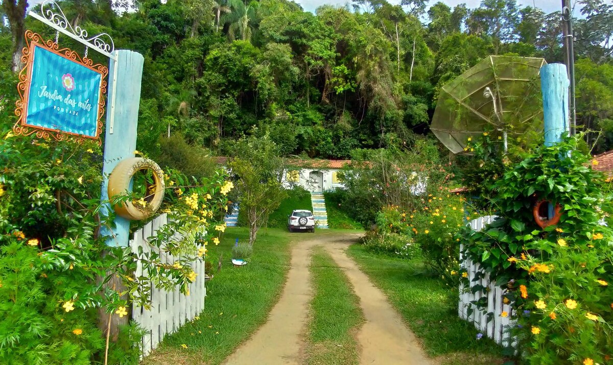 A charming entrance to the Jardim das Artes property is framed by lush greenery and blooming flowers. A dirt path leads to the main building, which is partially obscured by trees. Decorative elements like a sign and a large umbrella enhance the welcoming setting.