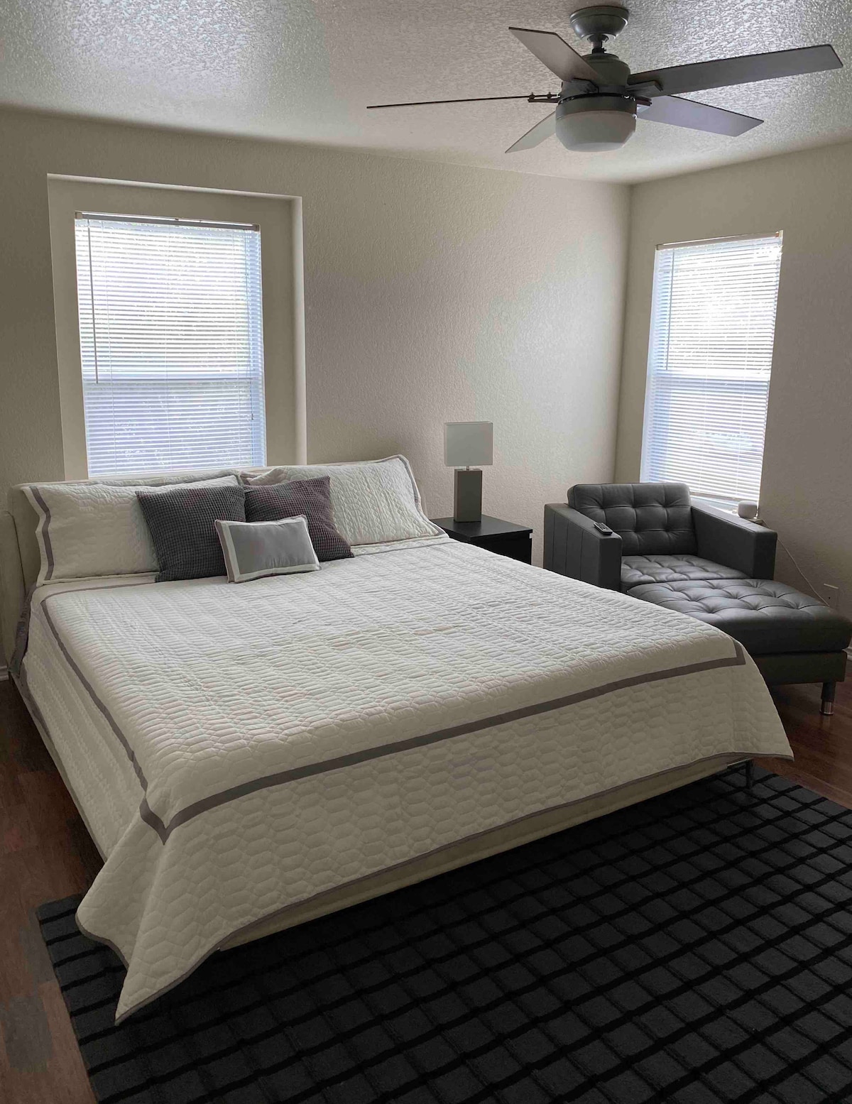 A spacious bedroom features a king bed dressed in a light quilt and decorative pillows. Natural light enters through two windows, complementing the modern furnishings, including a nightstand and a stylish chair. A dark area rug adds warmth to the hardwood floor.