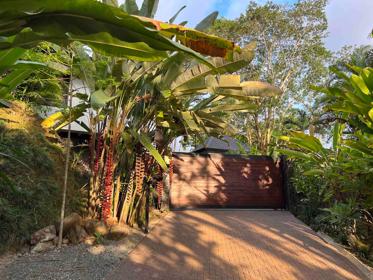 A serene entrance is framed by lush tropical foliage, including towering banana plants and vibrant red heliconias. A wooden gate stands closed, set against a backdrop of rolling hills and greenery, inviting guests to explore the accommodations within.