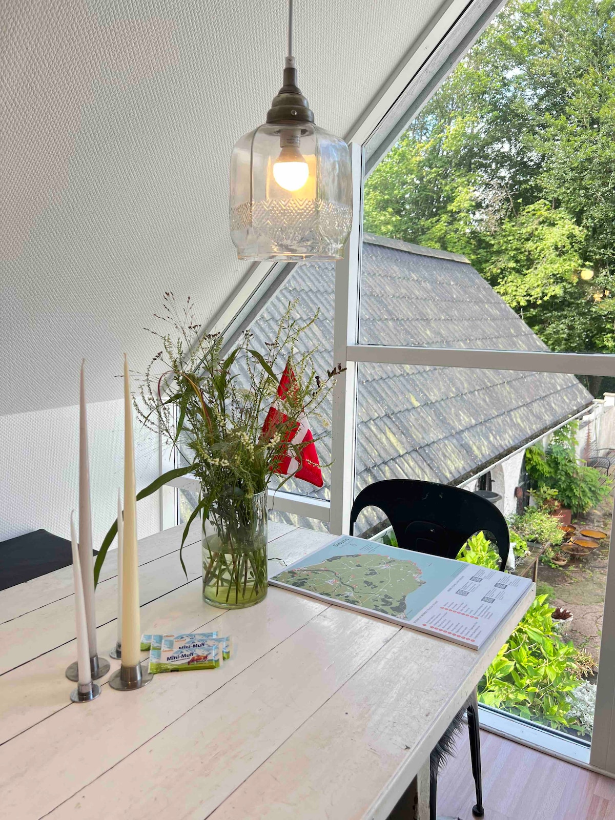 A dining table is positioned near a large glass window, offering views of the lush greenery outside. A vase of fresh flowers adorns the table, accompanied by two tall candles. A printed map is spread invitingly, and a black chair complements the setting.